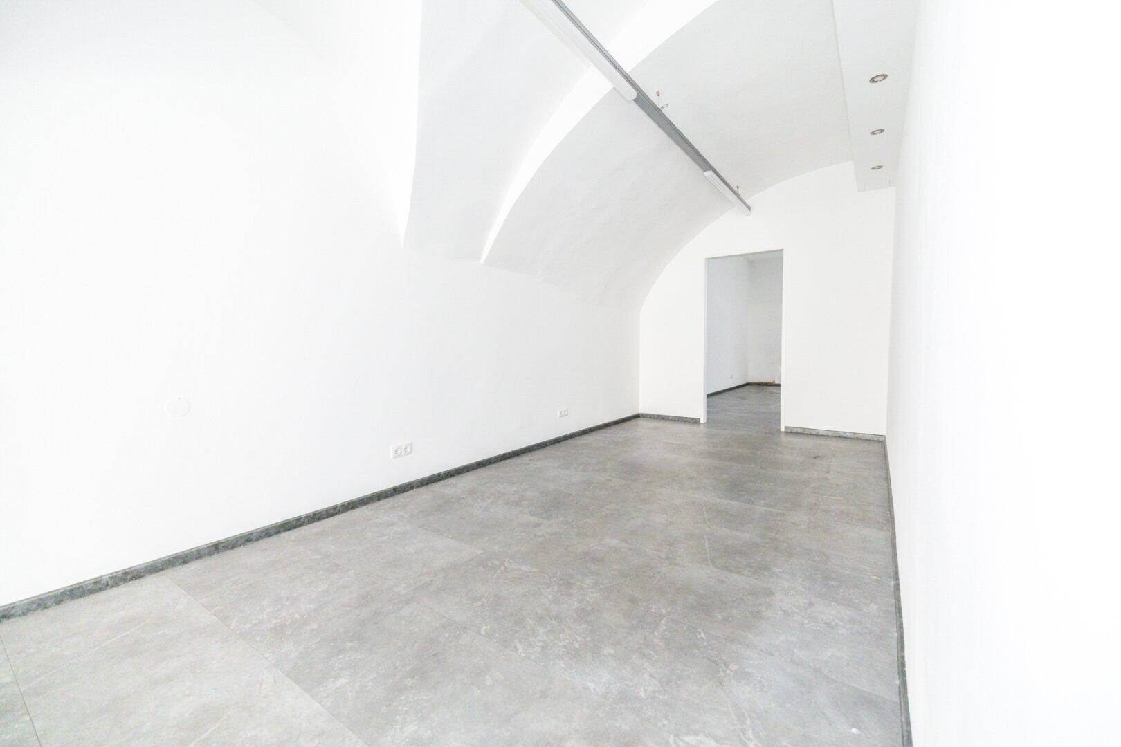 Weiterer Blick in den leeren Raum mit Gewölbedecken und Betonboden, der zu einem weiteren Bereich führt.