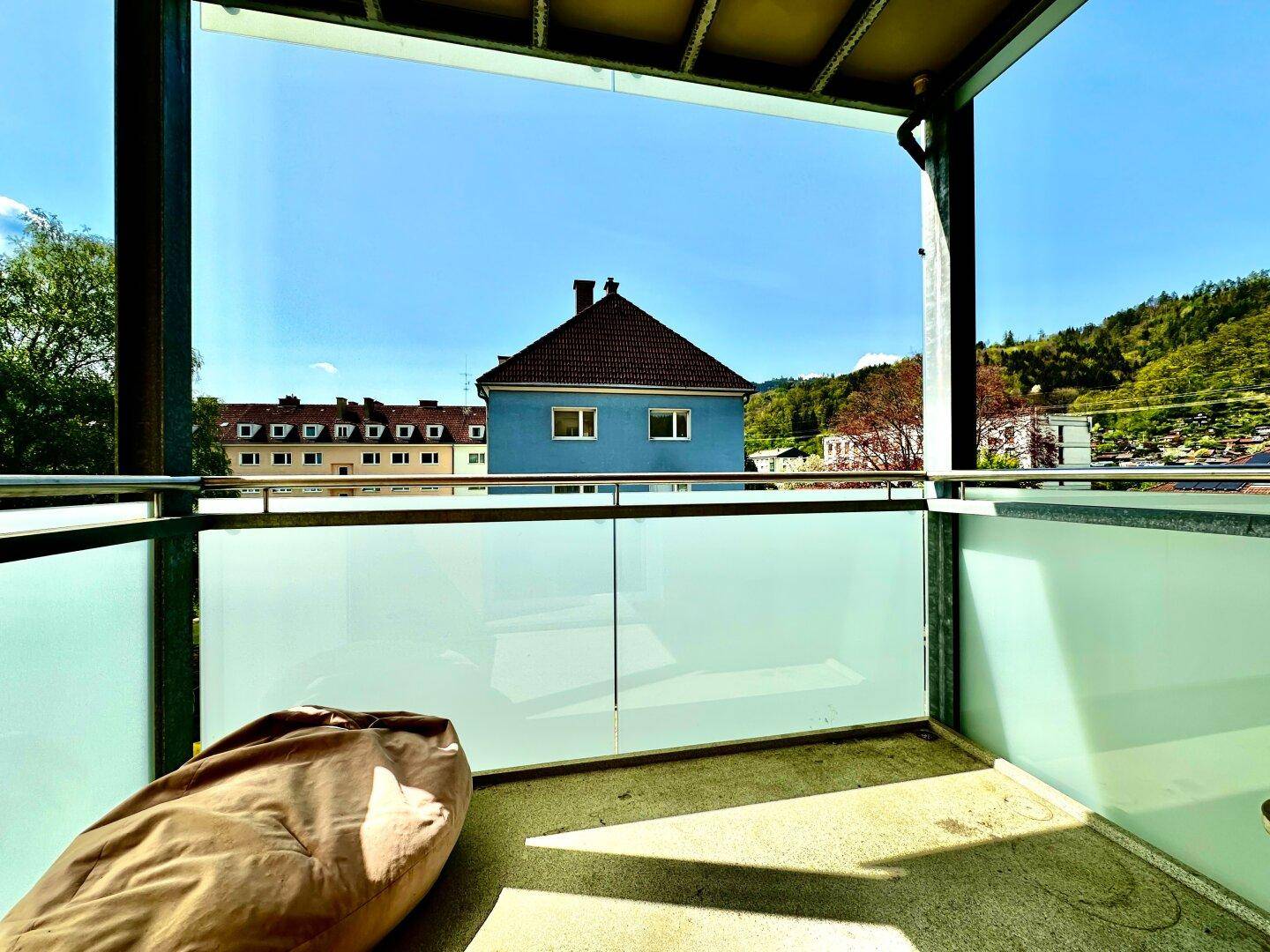 Heller Balkon mit Glasbrüstung und Blick auf die umliegenden Gebäude und grüne Hügel.