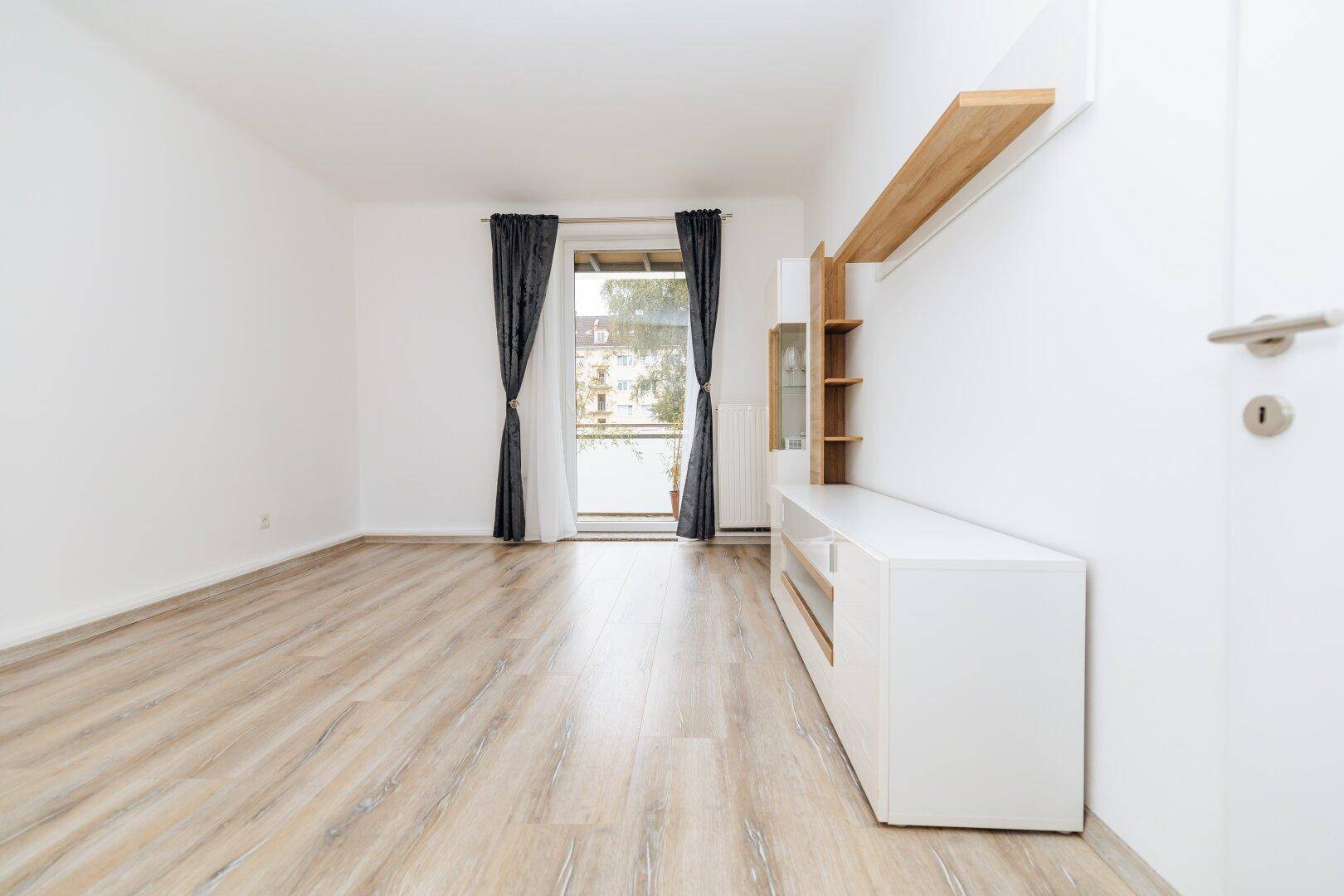 Helles Wohnzimmer mit Laminatboden, weiß gestrichenen Wänden und Zugang zu einem Balkon.