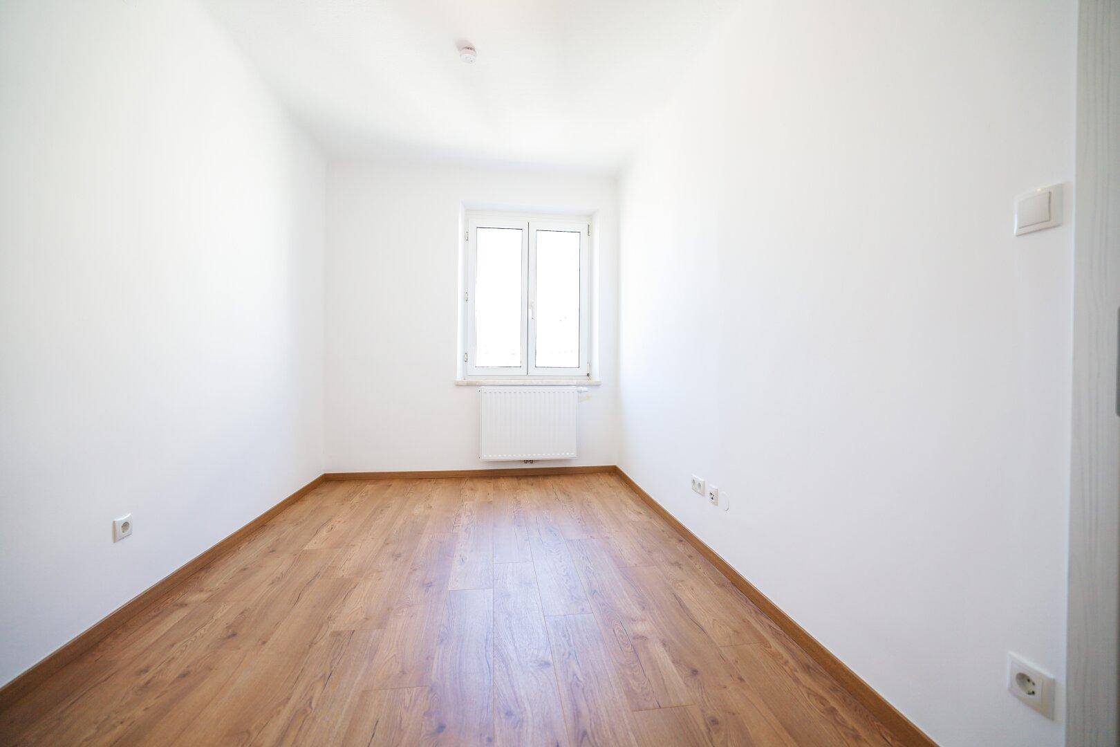 Schmaler, heller Raum mit Holzboden und einem Fenster, ideal als Schlaf- oder Arbeitszimmer.