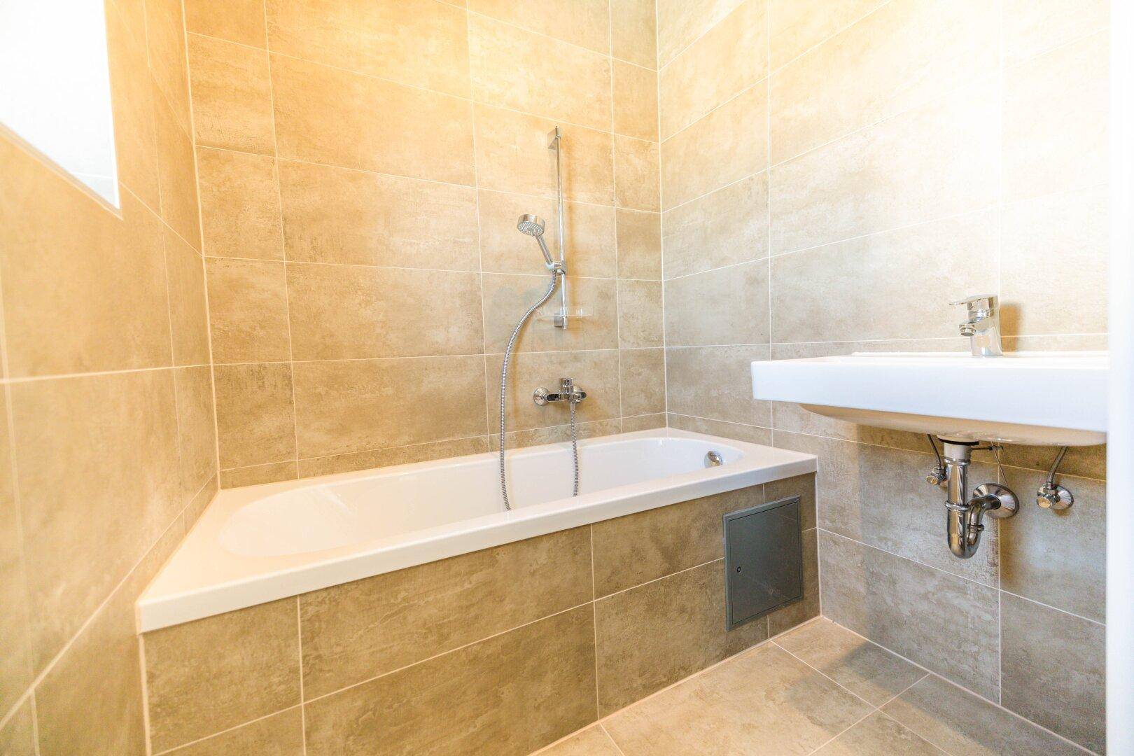 Modernes Badezimmer mit beigefarbenen Fliesen, einer Badewanne mit Duschfunktion und einem Waschbecken.