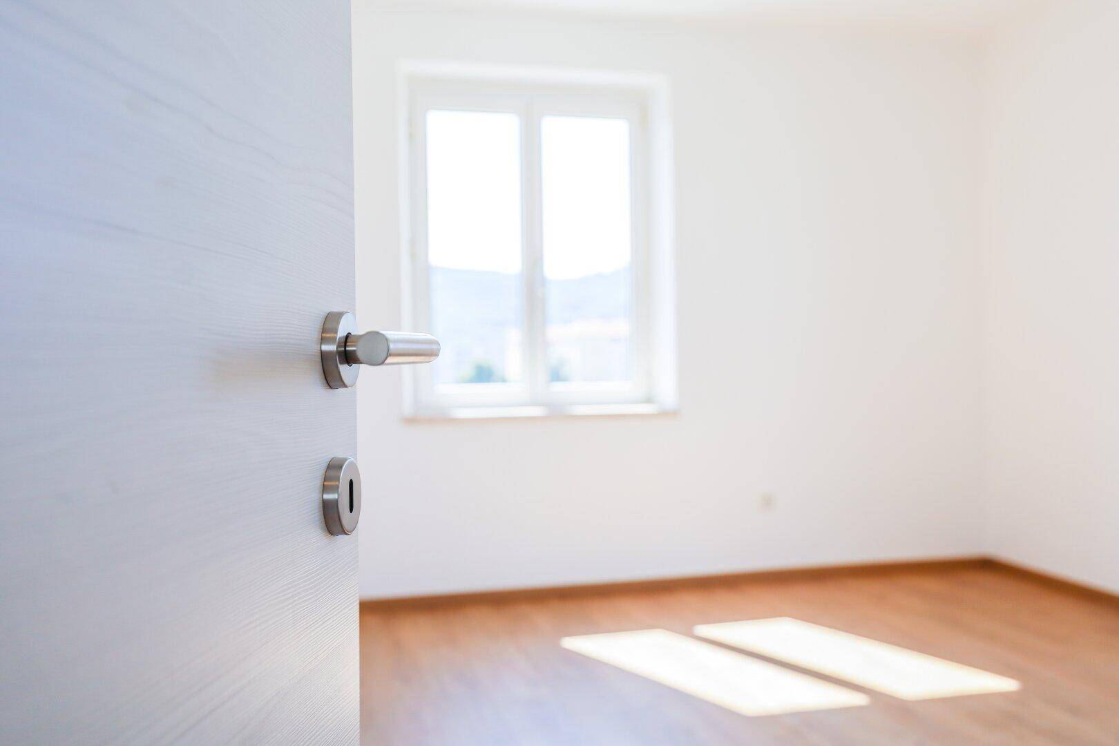 Offene Tür zu einem hellen, leeren Raum mit Holzboden und einem großen Fenster.