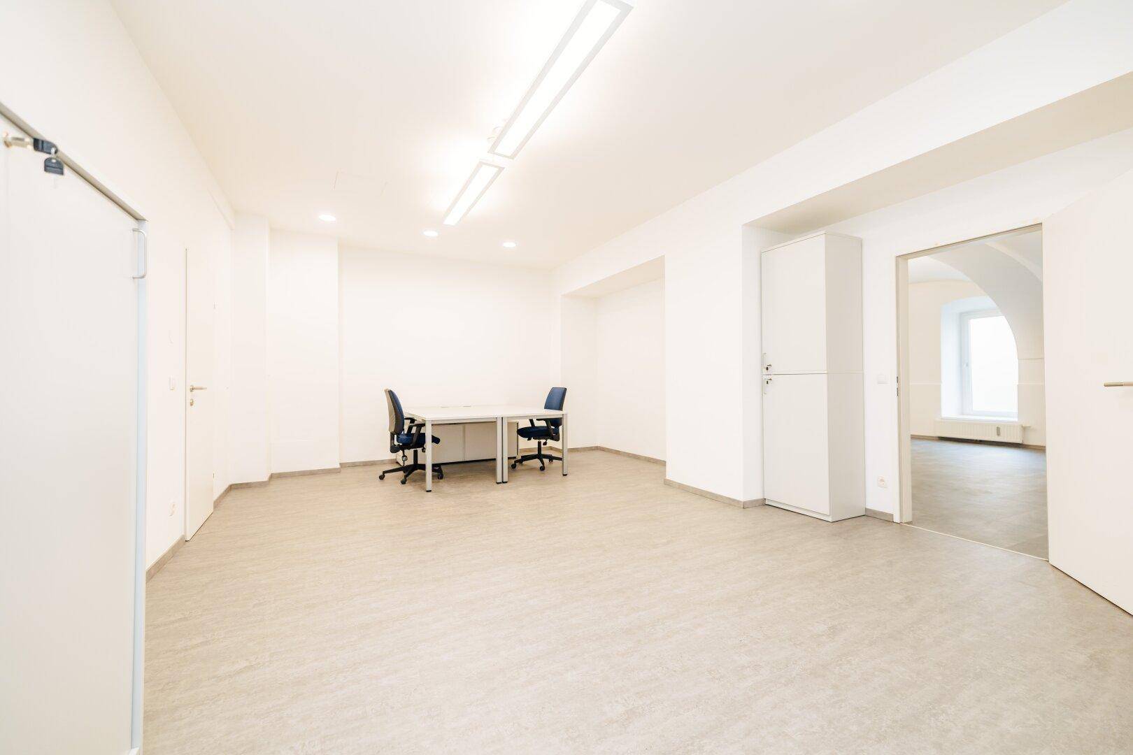 Geräumiges Büro mit Zugang zu einem weiteren Raum, hohen Decken und schlichten weißen Wänden.