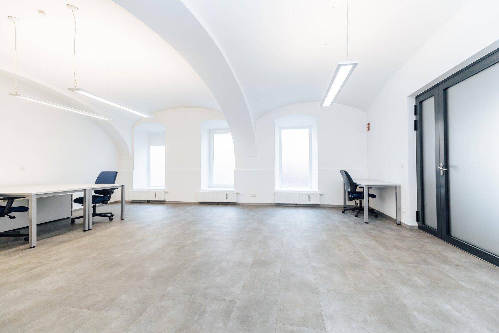 Heller Büroraum mit hohen Gewölbedecken, großen Fenstern und einer modernen Glastür.