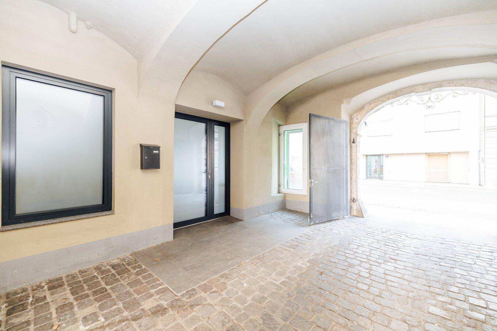 Historischer Eingangsbereich mit Gewölbebogen, Kopfsteinpflaster und moderner Glastür zum Gebäude.