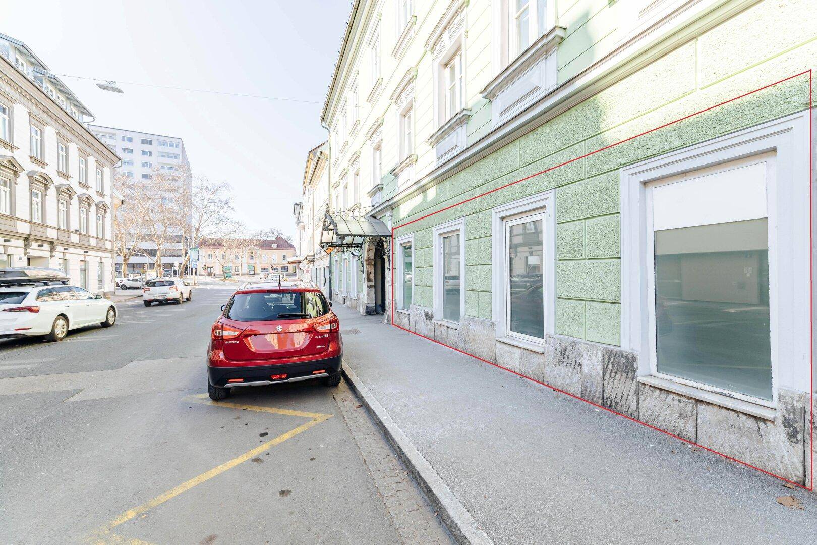 Historische Gebäudefassade an einer belebten Straße mit großen Fenstern und Parkmöglichkeiten.