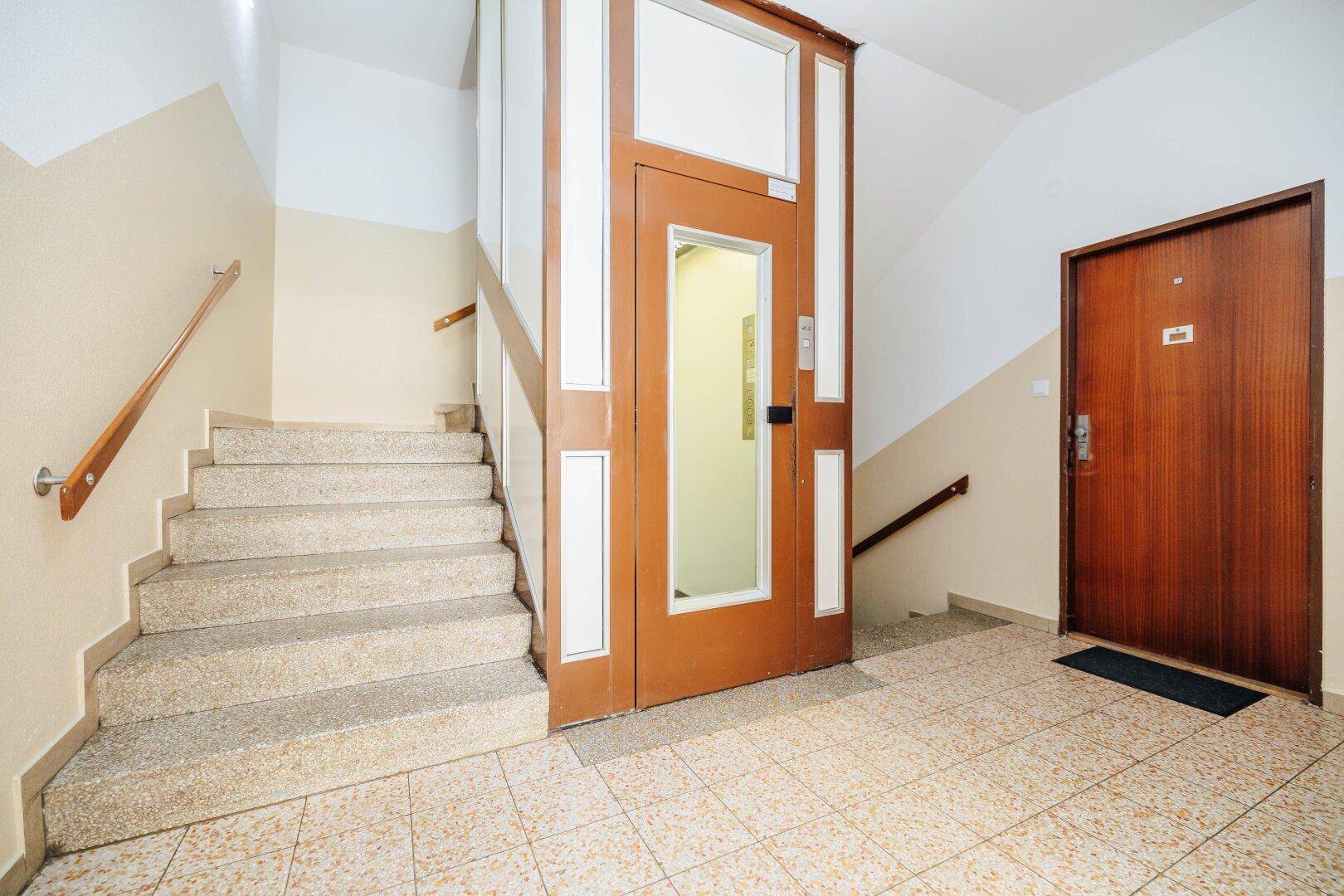 Eingangsbereich des Gebäudes mit Treppe, Aufzug und gefliestem Boden.