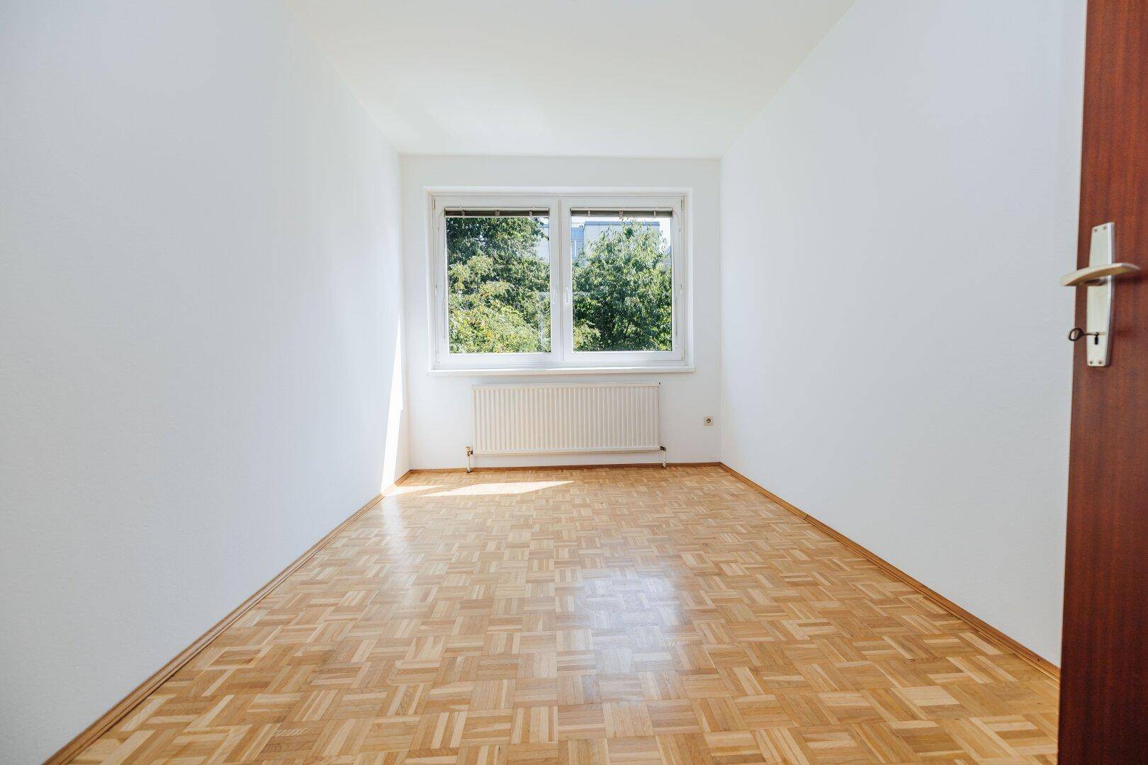 Helles Schlafzimmer mit Parkettboden und Fenster mit Blick auf Bäume.