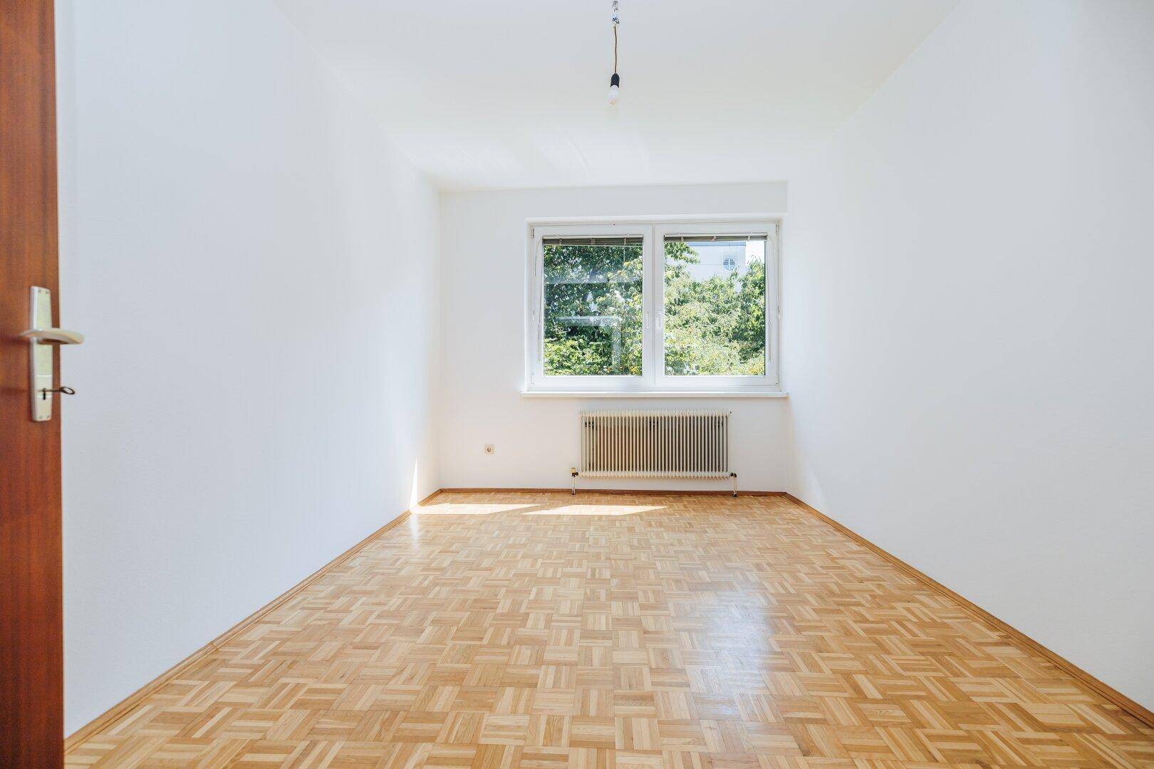 Leeres Schlafzimmer mit Parkettboden und Fenster zum grünen Außenbereich.