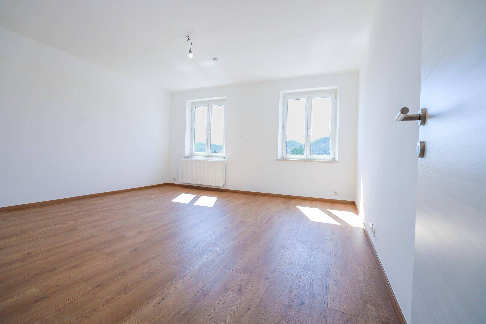 Geräumiger, heller Raum mit Holzboden und zwei Fenstern, die einen grünen Ausblick bieten.