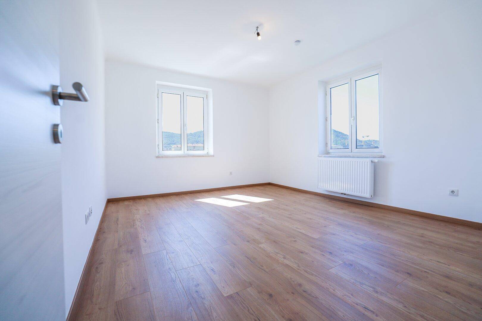 Lichtdurchfluteter Wohnraum mit Holzboden und weitem Blick ins Grüne durch zwei Fenster.