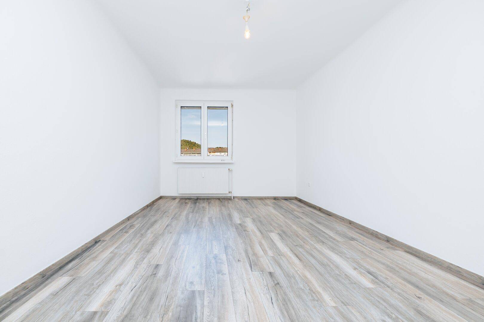 Helles, leeres Zimmer mit Fenster, Heizkörper und modernem Holzoptik-Bodenbelag.