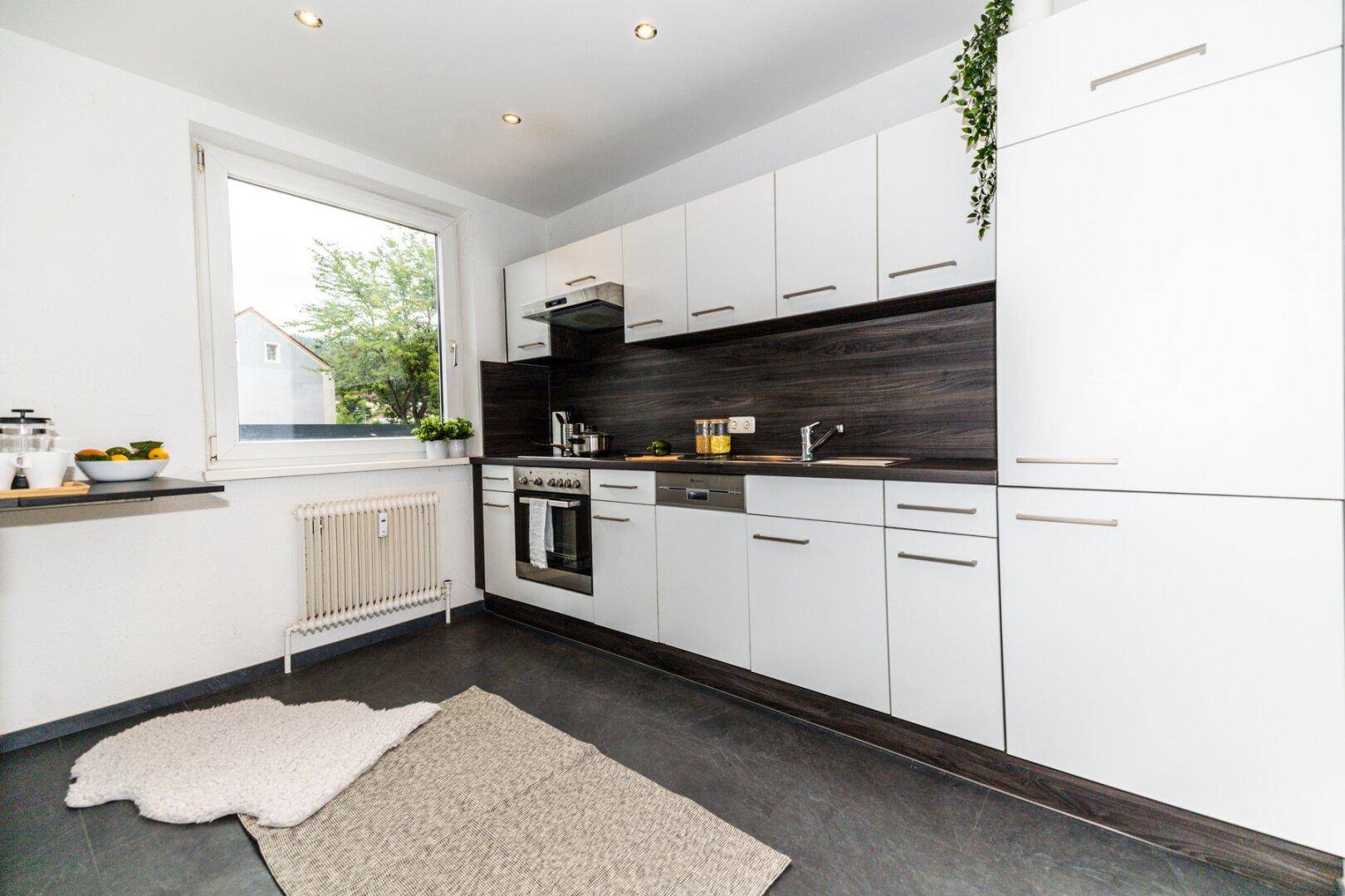 Moderne Einbauküche mit weißen Schränken, dunkler Arbeitsplatte und Fenster, ausgestattet mit Geräten.