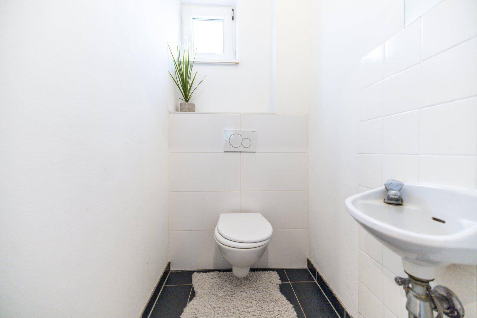 Separates WC mit wandhängender Toilette, kleinem Waschbecken, weißen Fliesen und Fenster.