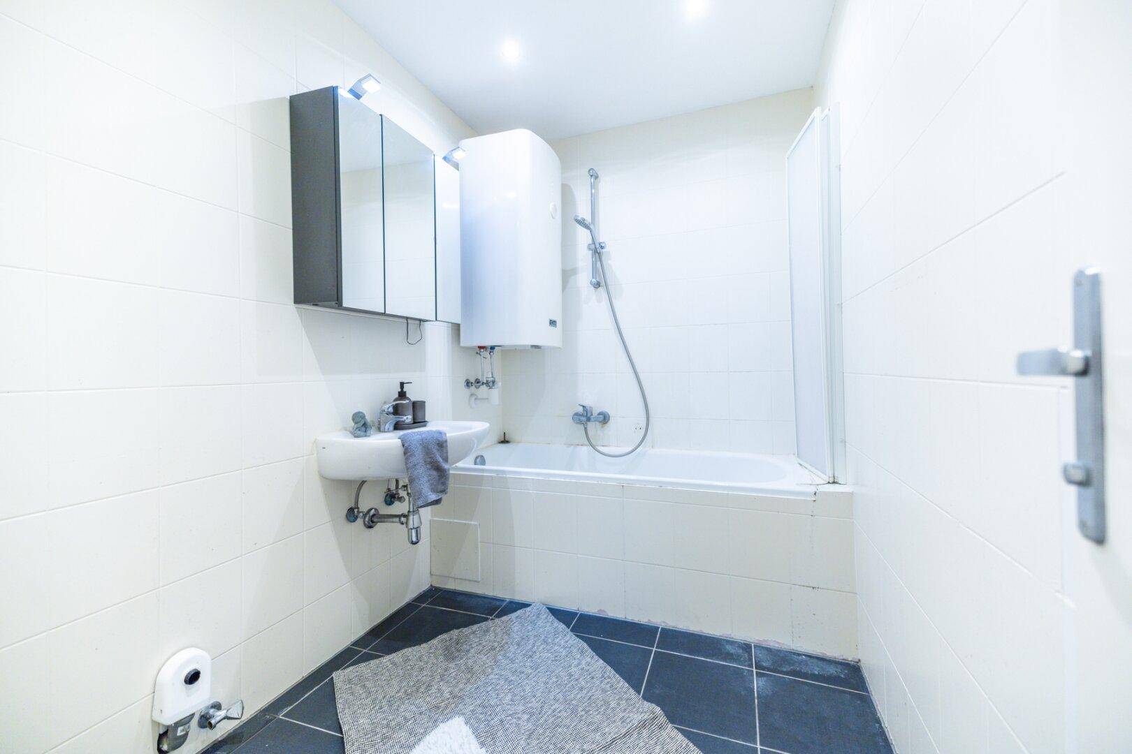 Badezimmer mit weißen Fliesen, dunklem Boden, Badewanne mit Dusche, Waschbecken und Spiegelschrank.