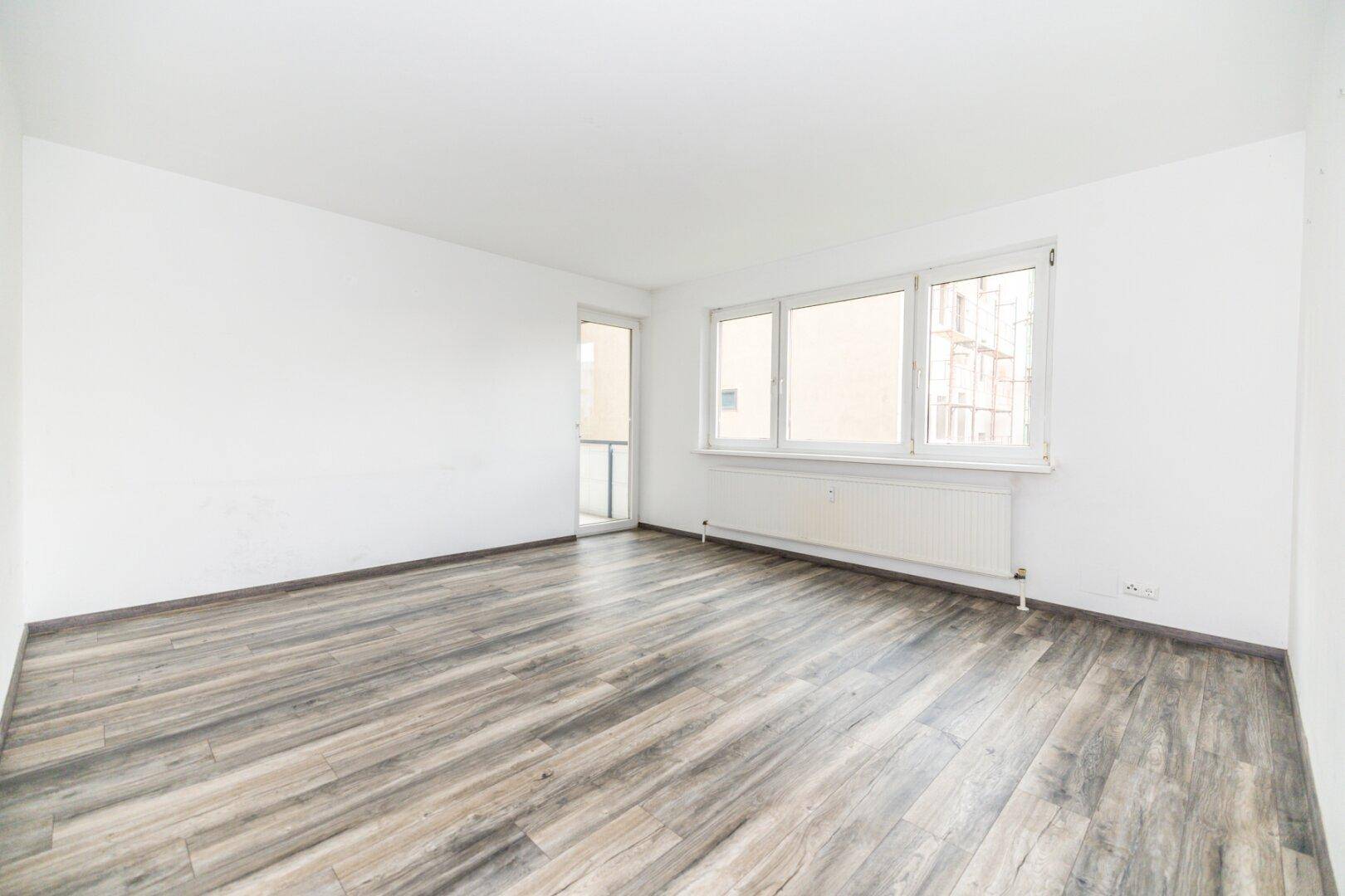 Geräumiges Wohnzimmer mit Laminatboden, weißen Wänden, großen Fenstern und Balkonzugang.