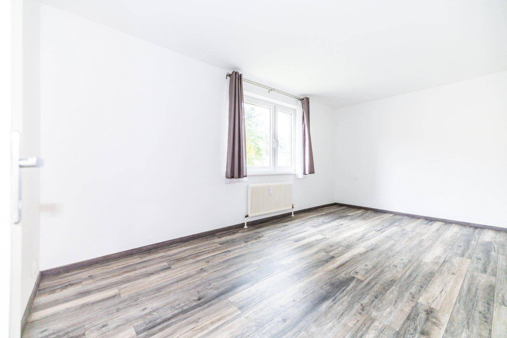 Geräumiges Schlafzimmer mit Laminatboden, weißen Wänden und einem Fenster mit Vorhängen.