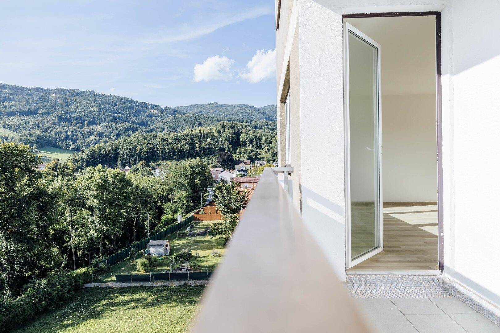 Sonniger Balkon mit Blick ins Grüne und auf bewaldete Hügel, mit Zugang zu einem hellen Raum.