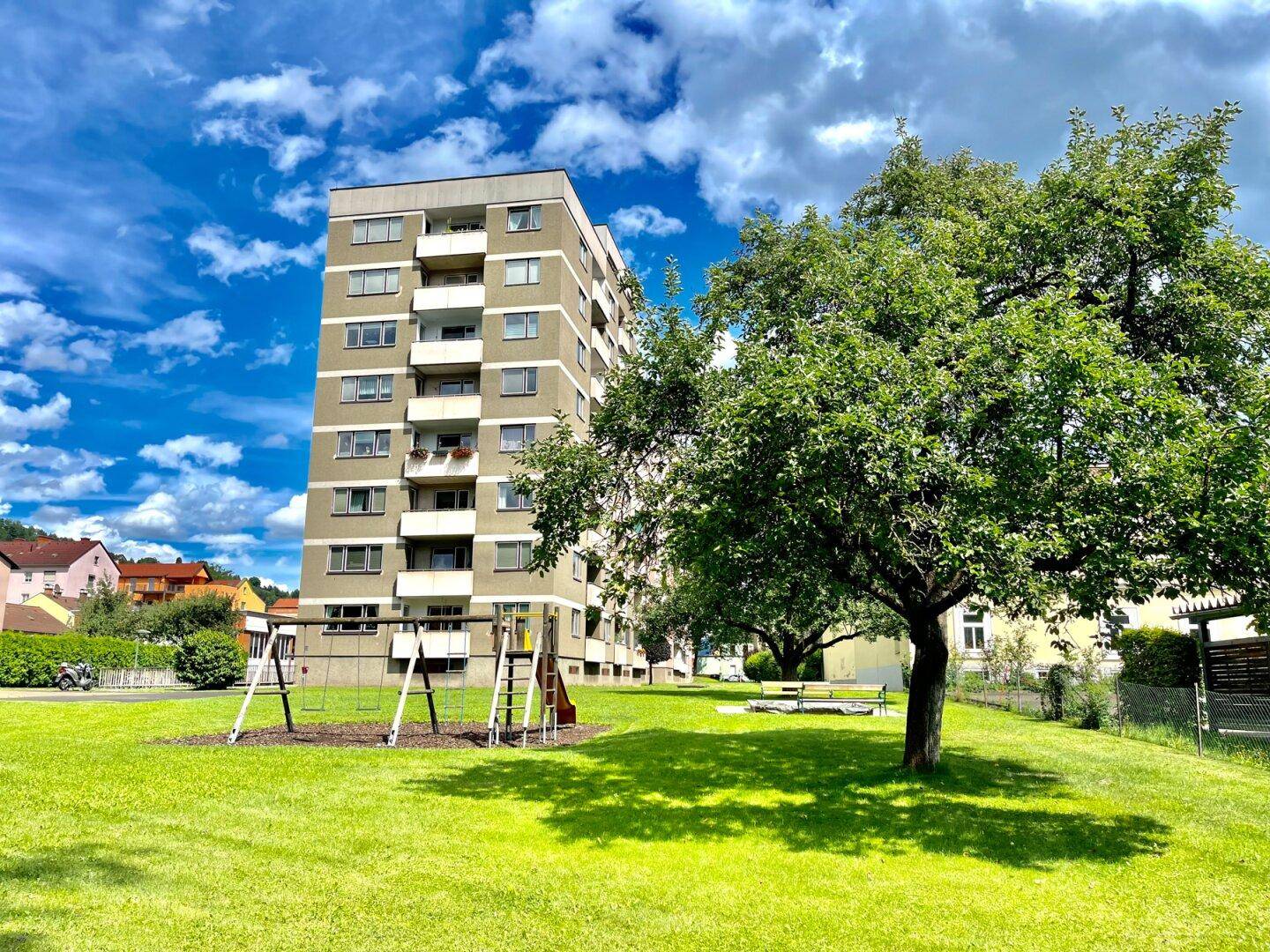 Mehrfamilienhaus mit großzügiger Grünfläche, Kinderspielplatz und altem Baumbestand im Vordergrund.