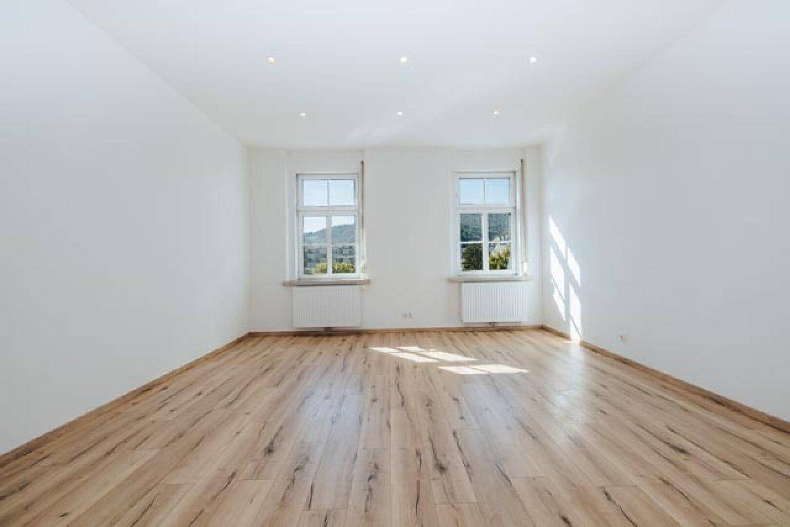 Helles, leeres Zimmer mit Laminatboden, weißen Wänden und zwei Fenstern mit Blick ins Grüne.