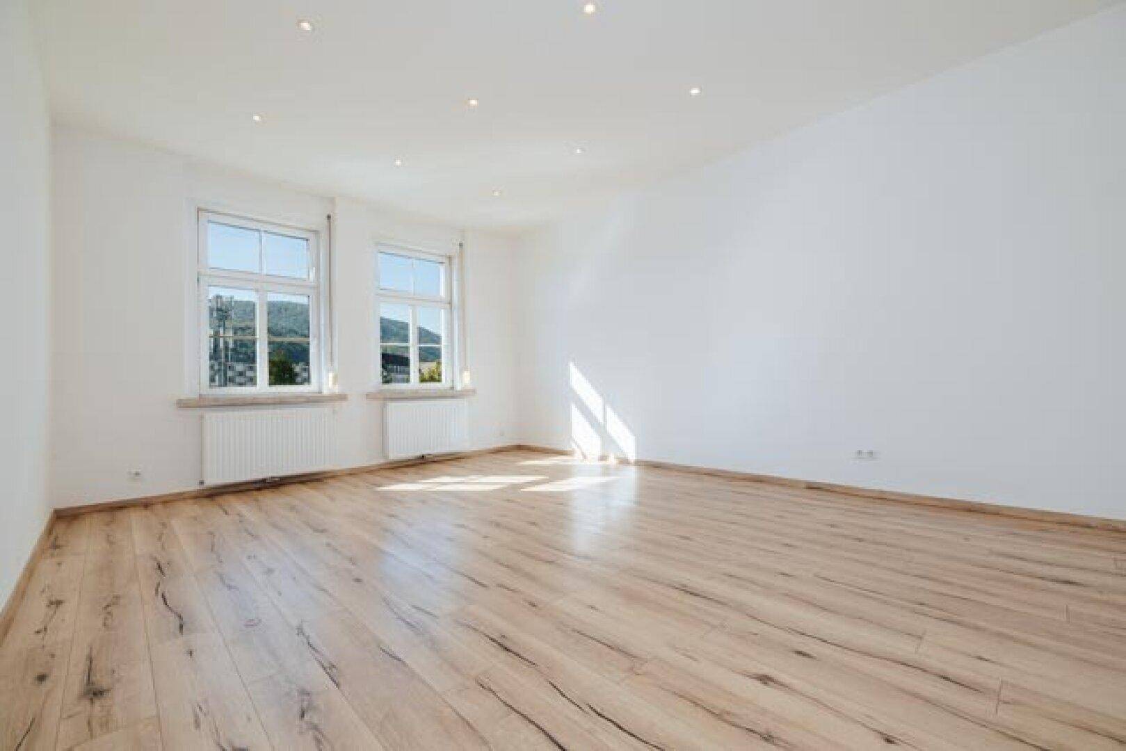 Lichtdurchfluteter Raum mit hellem Laminatboden, weißen Wänden und Fenstern, die viel Tageslicht hereinlassen.