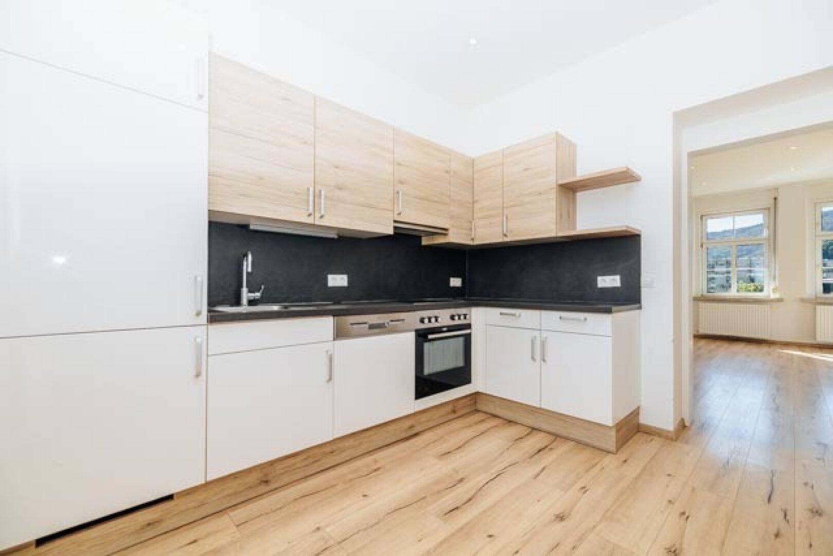 Geräumige Einbauküche mit Holz- und weißen Schränken, schwarzer Arbeitsplatte und offenem Übergang zu einem weiteren Raum.