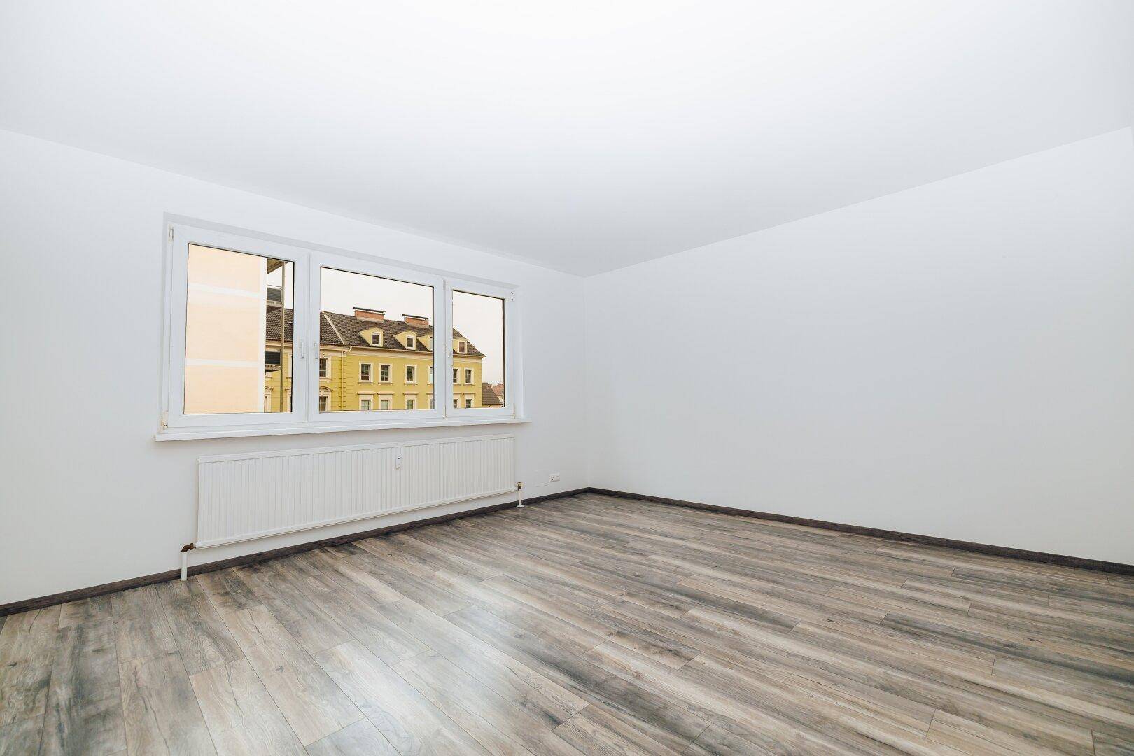 Heller, leerer Raum mit weißen Wänden, modernem Laminatboden und Blick auf ein gegenüberliegendes Gebäude.