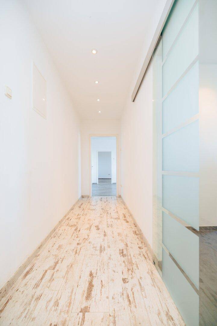 Langer, heller Flur mit hellem Holzboden, Einbaustrahlern und einer Glasschiebetür.