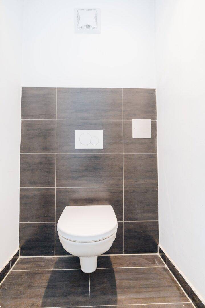 Modernes WC mit wandhängender Toilette, dunklen Fliesen und Abluftsystem.