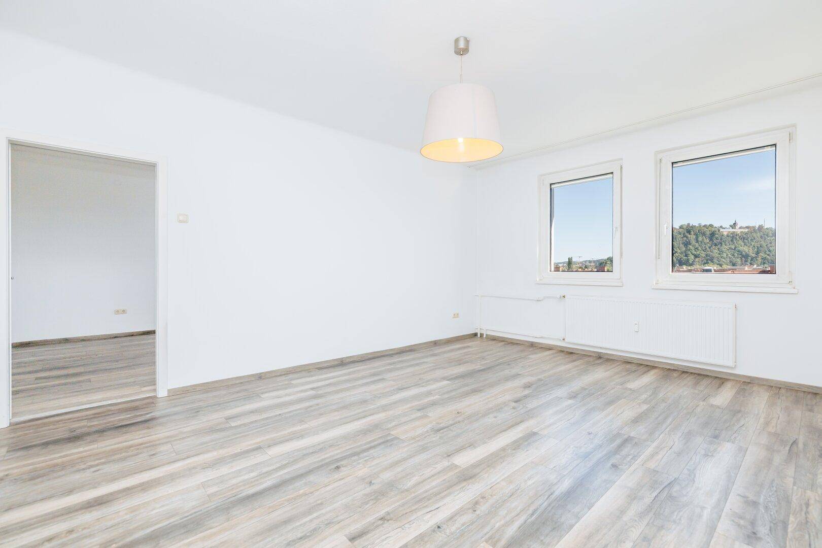 Helles, leeres Zimmer mit modernem Laminatboden und zwei Fenstern mit Ausblick.