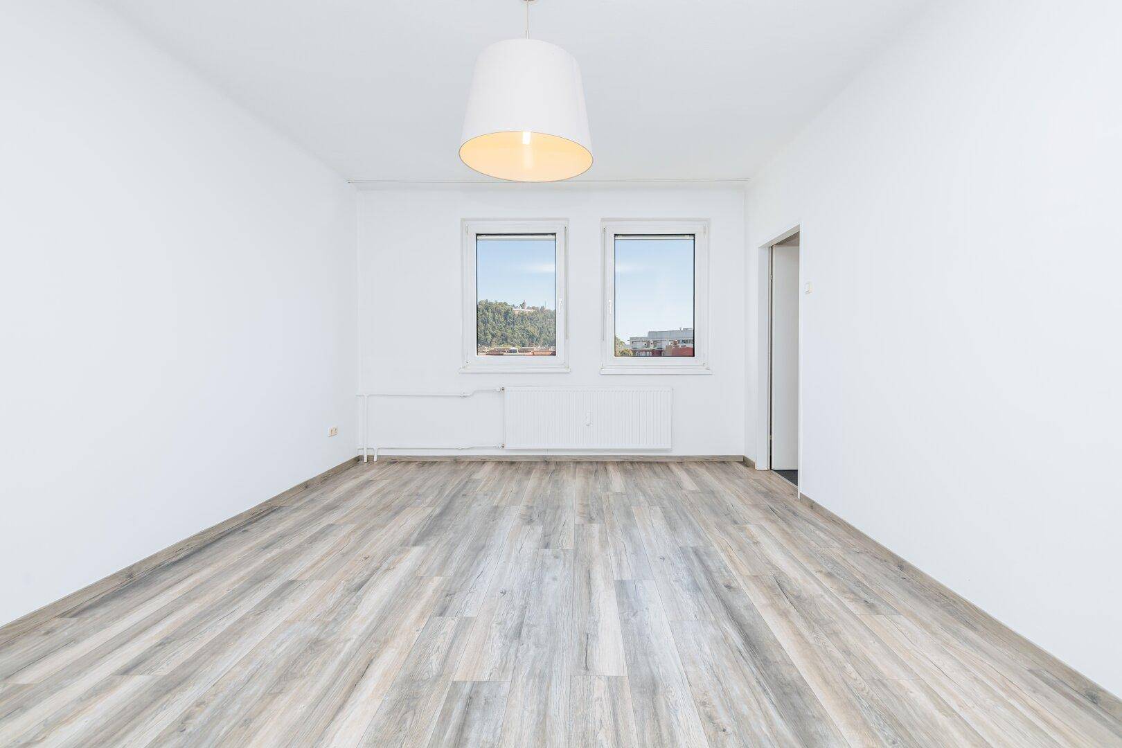 Geräumiges, unmöbliertes Zimmer mit hellem Laminatboden und viel Tageslicht durch die Fenster.
