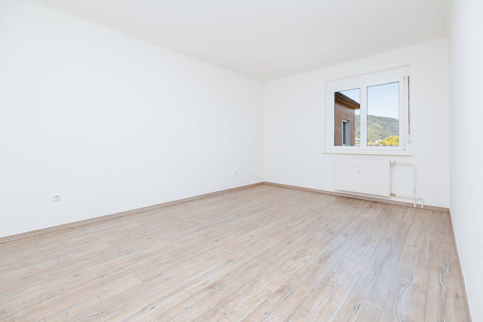 Ruhiges Schlafzimmer mit hellem Laminatboden und Fenster, das einen Blick ins Grüne bietet.