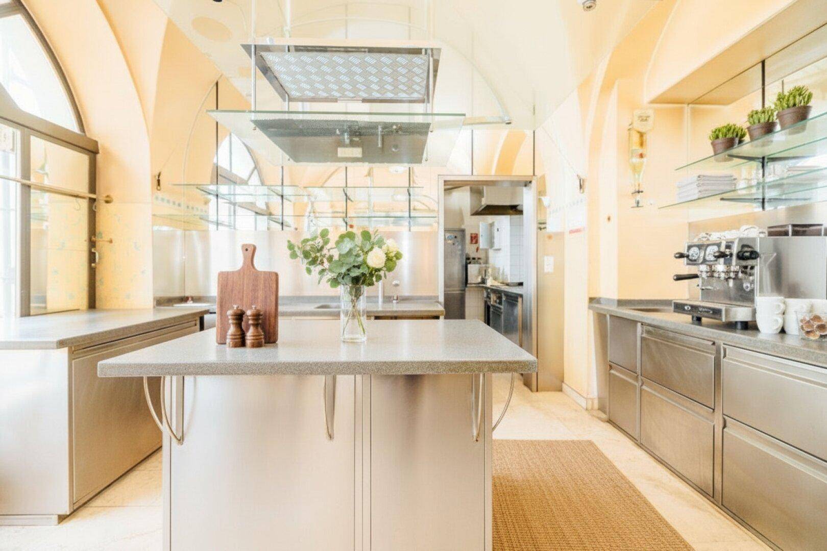 Professionelle Edelstahlküche mit Kochinsel, Dunstabzugshauben und Kaffeemaschine in einem gewölbten Raum.