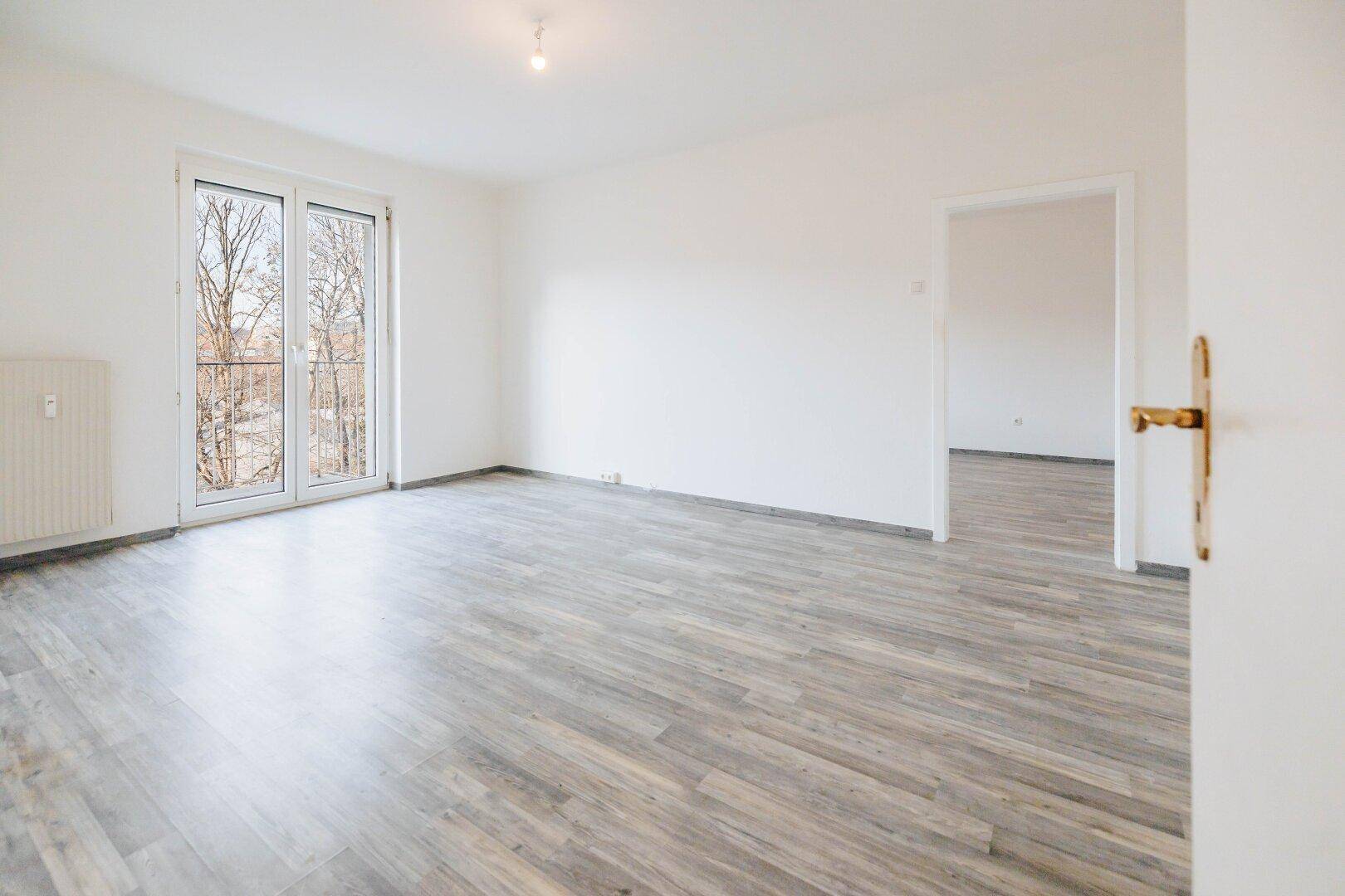 Geräumiger, heller Wohnbereich mit Laminatboden und Zugang zum Balkon sowie einem weiteren Zimmer.