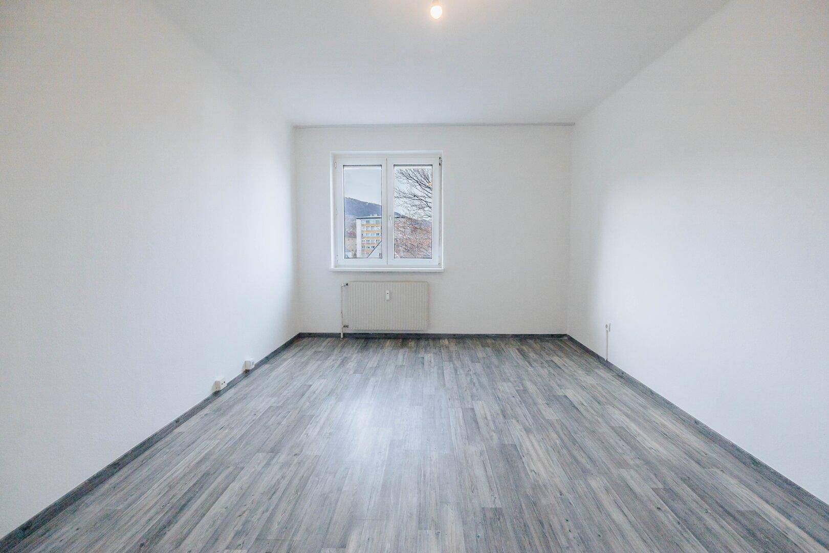 Schlichtes Zimmer mit weiß gestrichenen Wänden, Laminatboden und einem Fenster mit Ausblick.