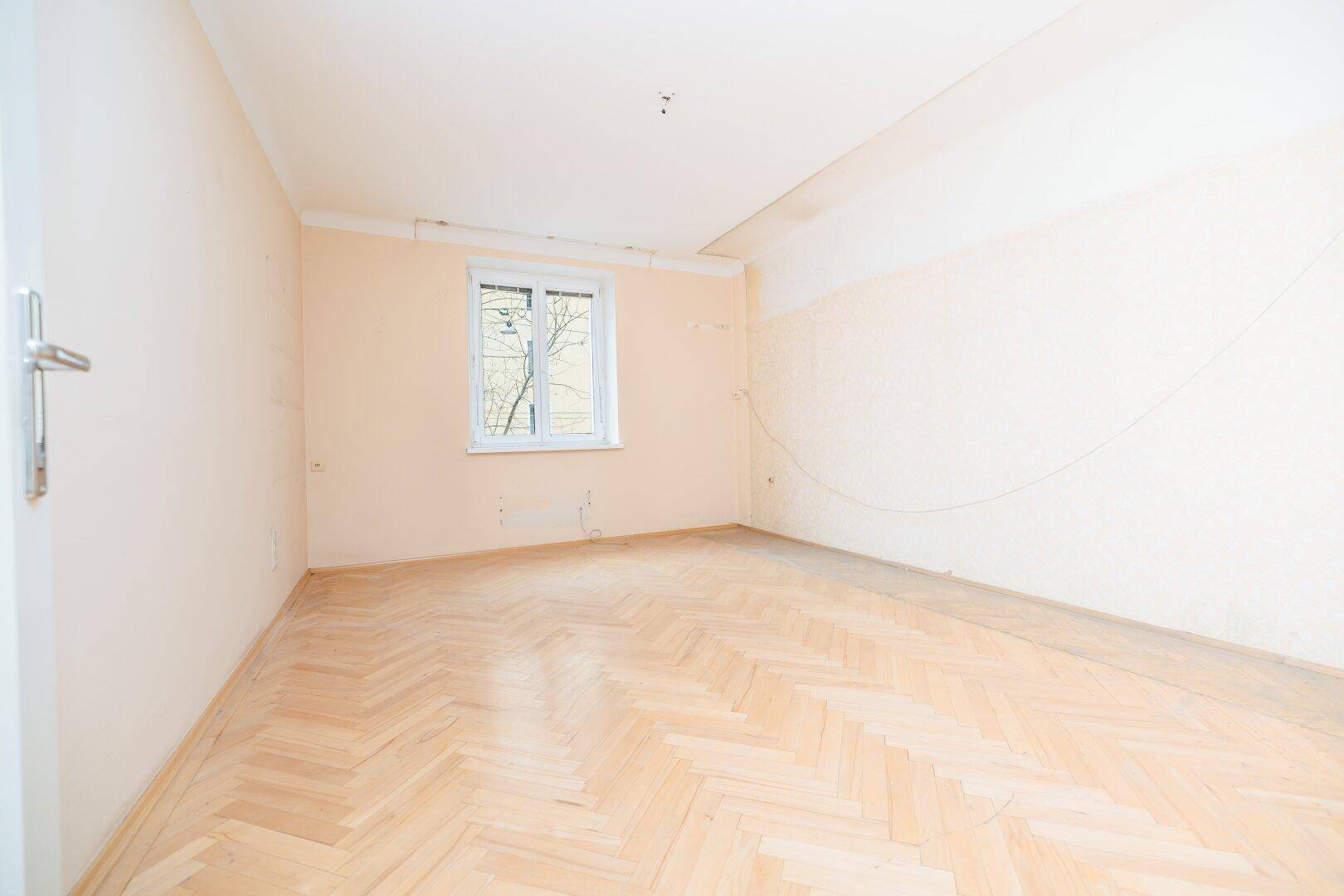 Leeres Zimmer mit hellem Fischgrätparkettboden und einem Fenster, bereit zur Renovierung.