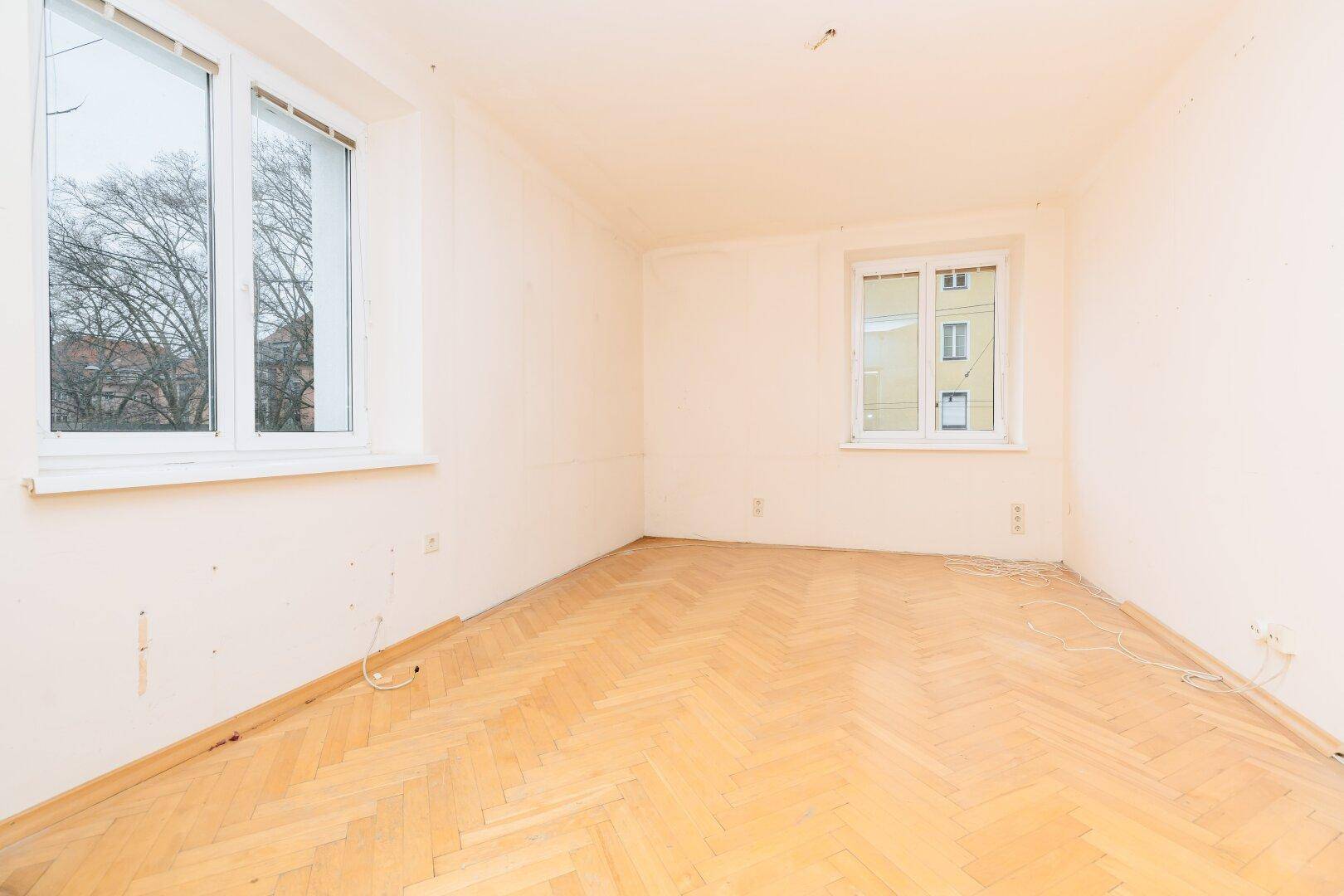 Leeres Zimmer mit hellem Fischgrätparkettboden und großem Fenster mit Blick ins Grüne.