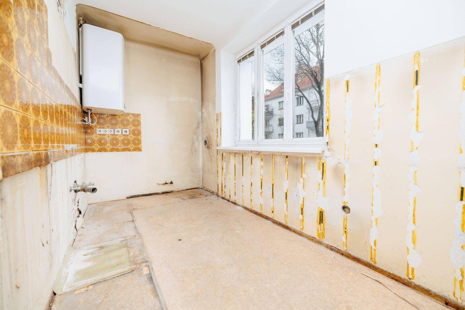 Leerer Küchenraum mit Fenster, Wasserboiler und Spuren alter Fliesen, bereit zur Renovierung.