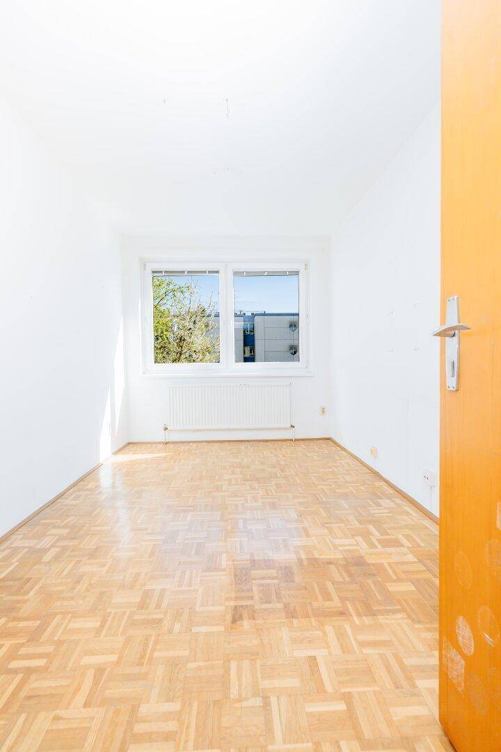 Helles Zimmer mit Parkettboden, weißen Wänden und Fensterblick ins Grüne.