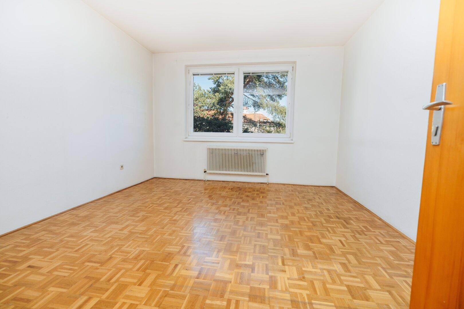 Ruhiges Zimmer mit Parkettboden, weißen Wänden und Blick auf Bäume.