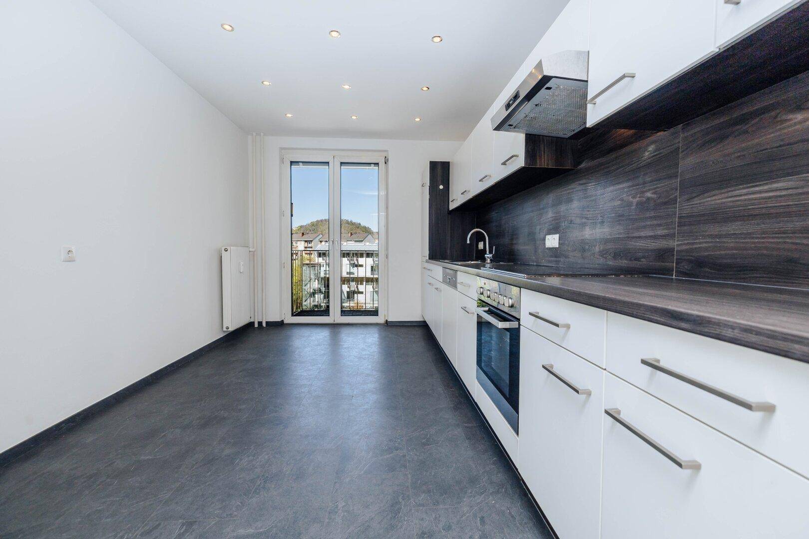 Moderne Einbauküche mit weißen Schränken, dunkler Arbeitsplatte und Zugang zu einem Balkon.