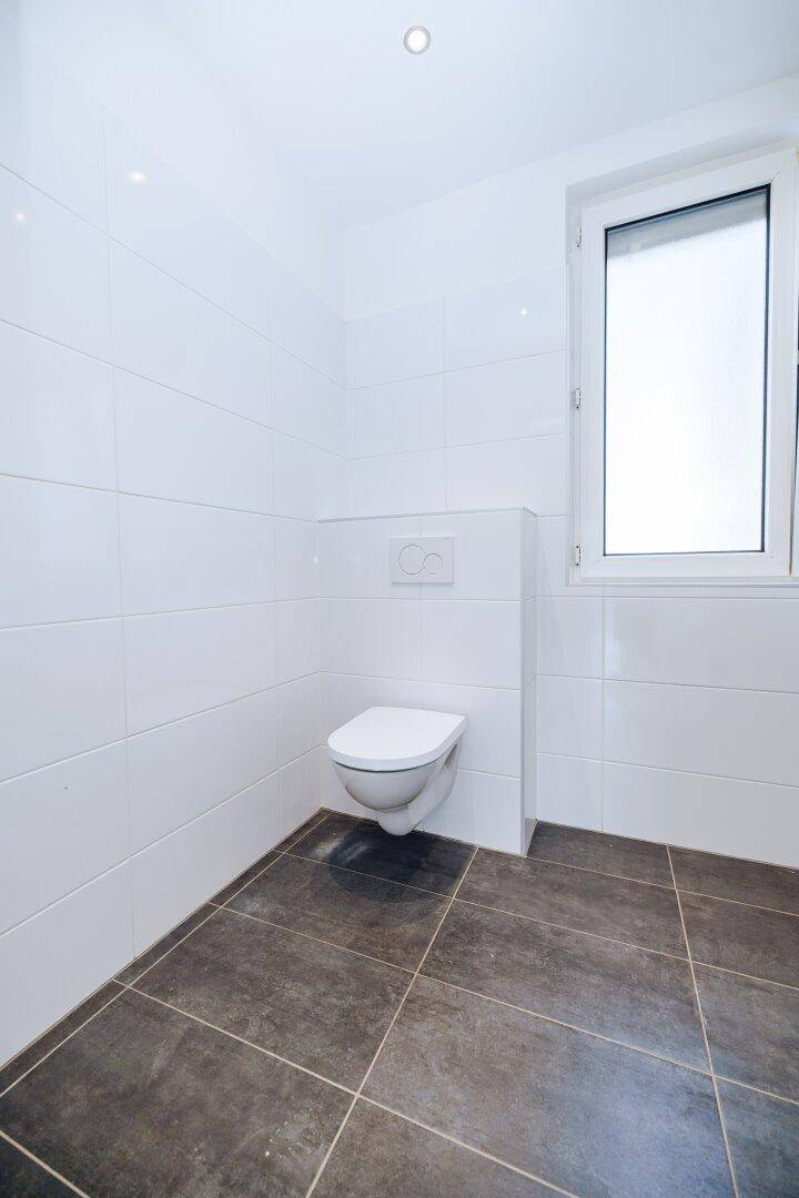 Separates WC mit wandhängender Toilette, Fenster und modernen Fliesen in Weiß und Grau.