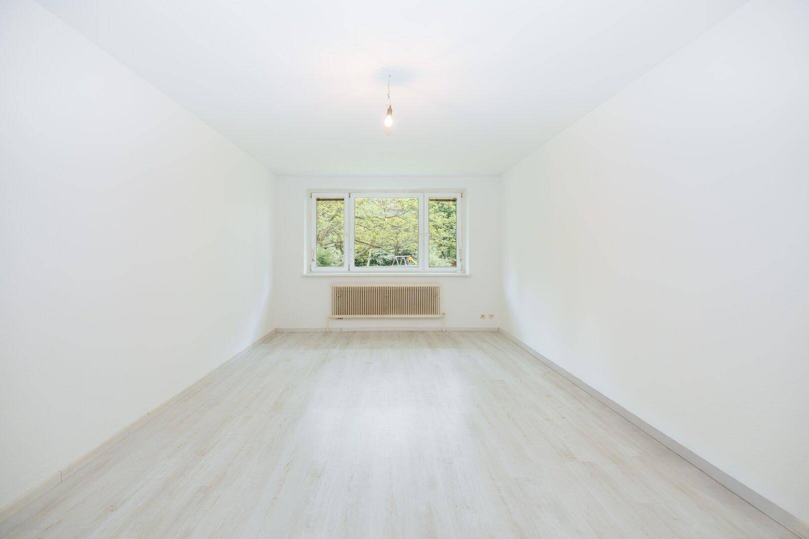 Geräumiger, leerer Raum mit hellem Laminatboden und großen Fenstern mit Blick ins Grüne.