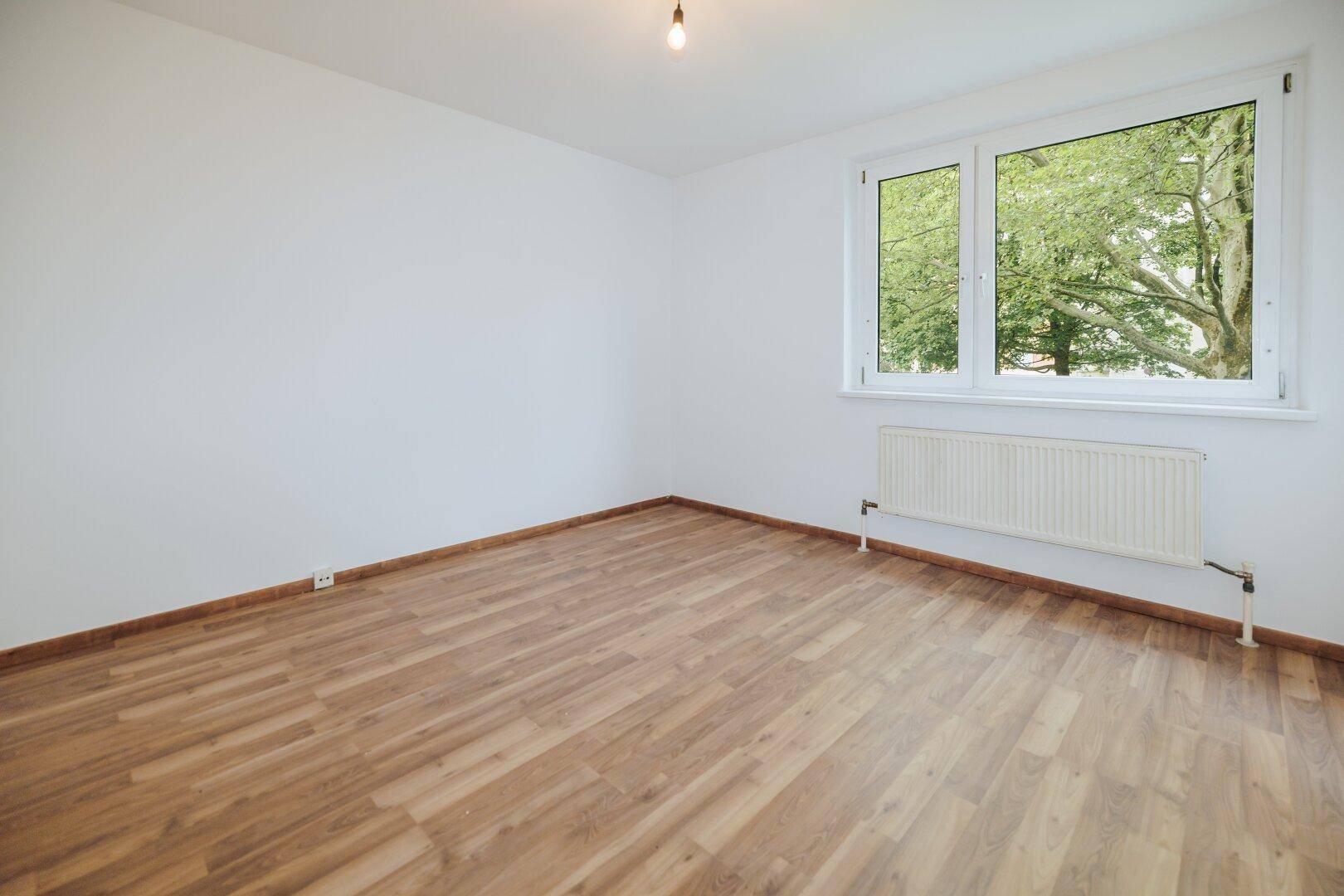 Heller, leerer Raum mit Laminatboden und einem Fenster, das einen Blick auf Bäume bietet.