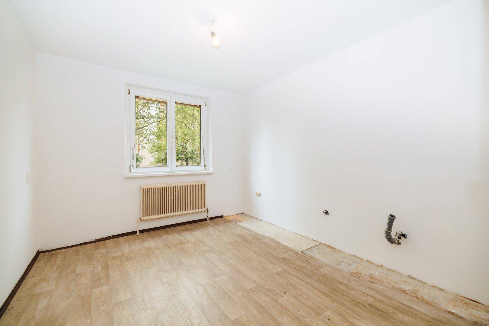 Leerer Raum mit Laminatboden, weißen Wänden und sichtbaren Wasseranschlüssen für eine mögliche Küche.
