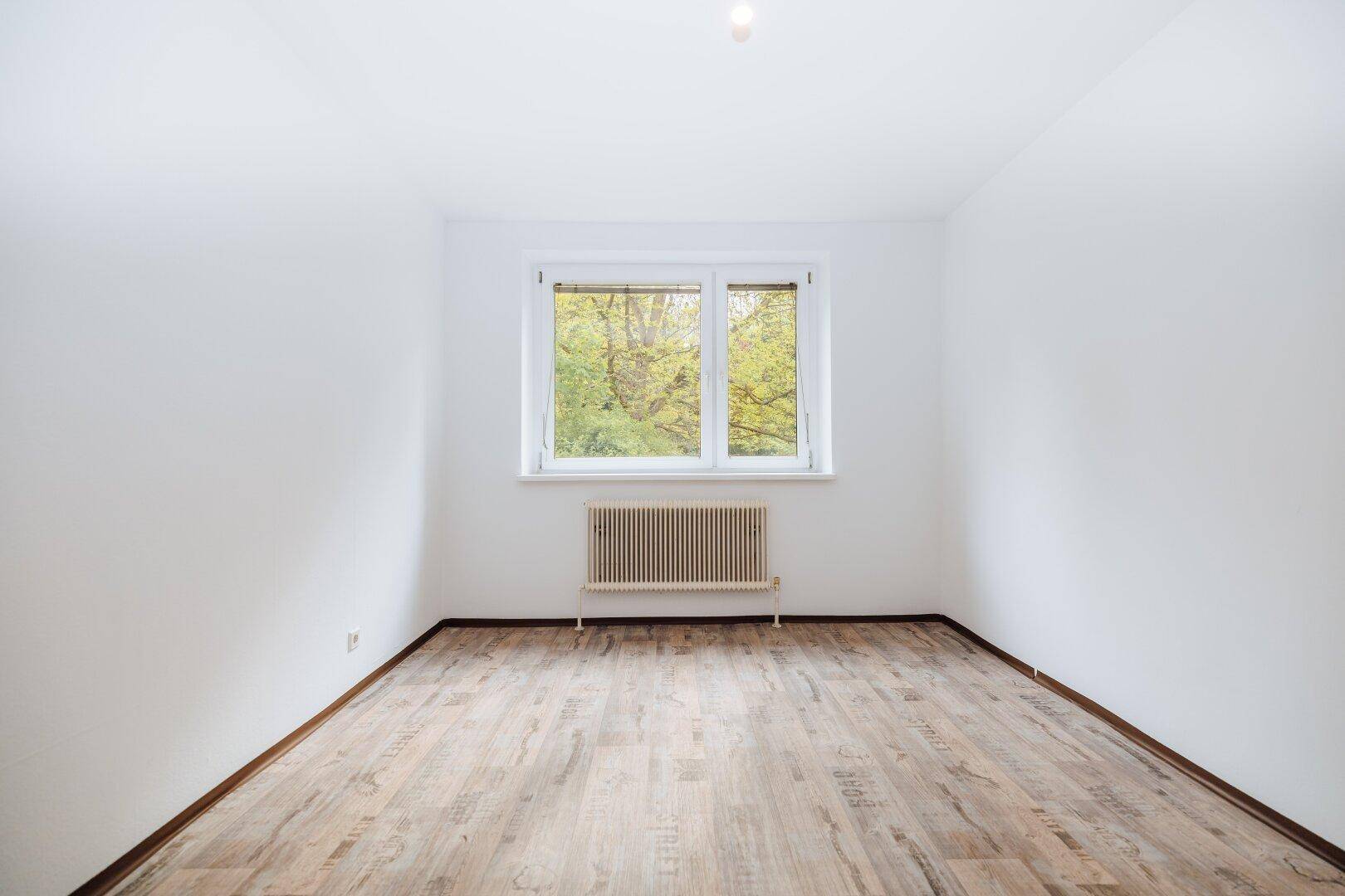 Leerer Raum mit Laminatboden, weißen Wänden und einem Fenster mit Blick auf die umliegenden Bäume.