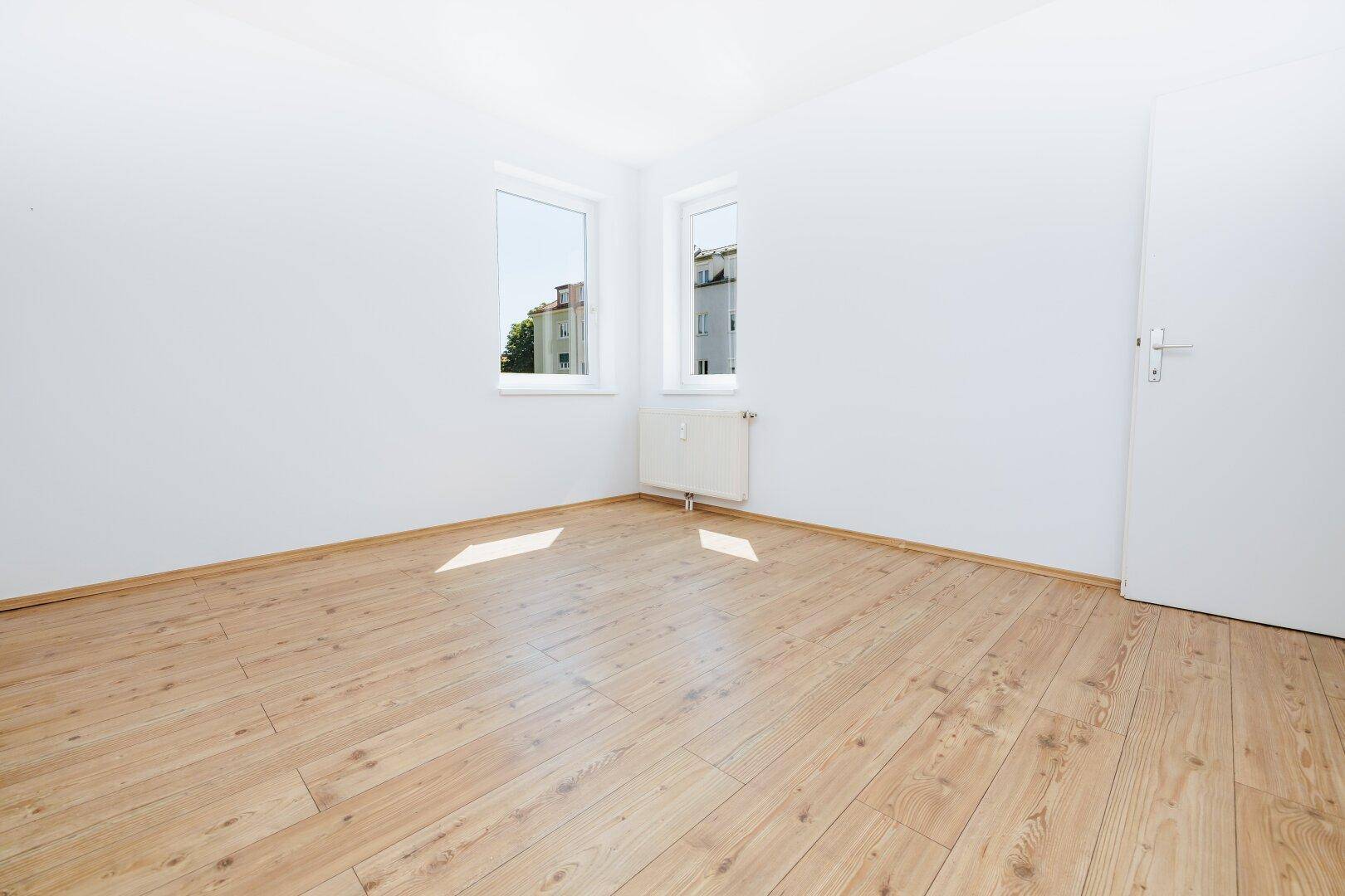 Helles, leeres Zimmer mit weißen Wänden, Holzdielenboden und zwei Fenstern mit Blick ins Grüne.