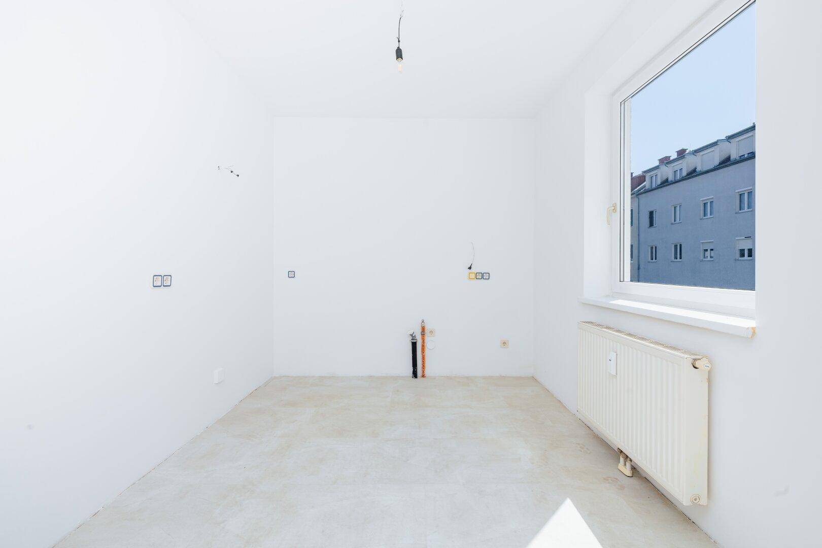 Leerer Küchenraum mit Estrichboden, weißen Wänden und großem Fenster mit Stadtblick.
