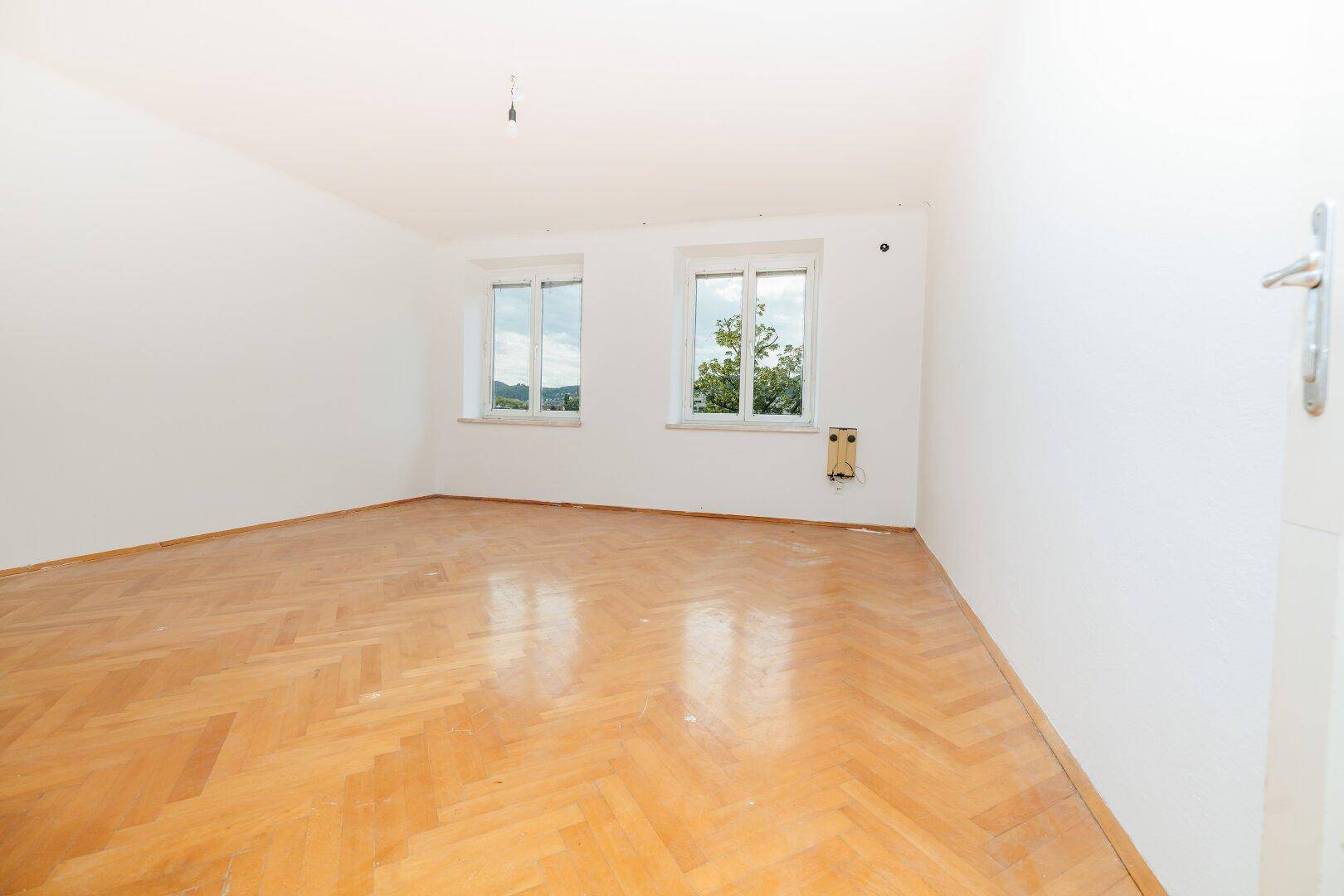 Geräumiges, leeres Zimmer mit Fischgrätparkettboden und zwei Fenstern für natürliche Belichtung.