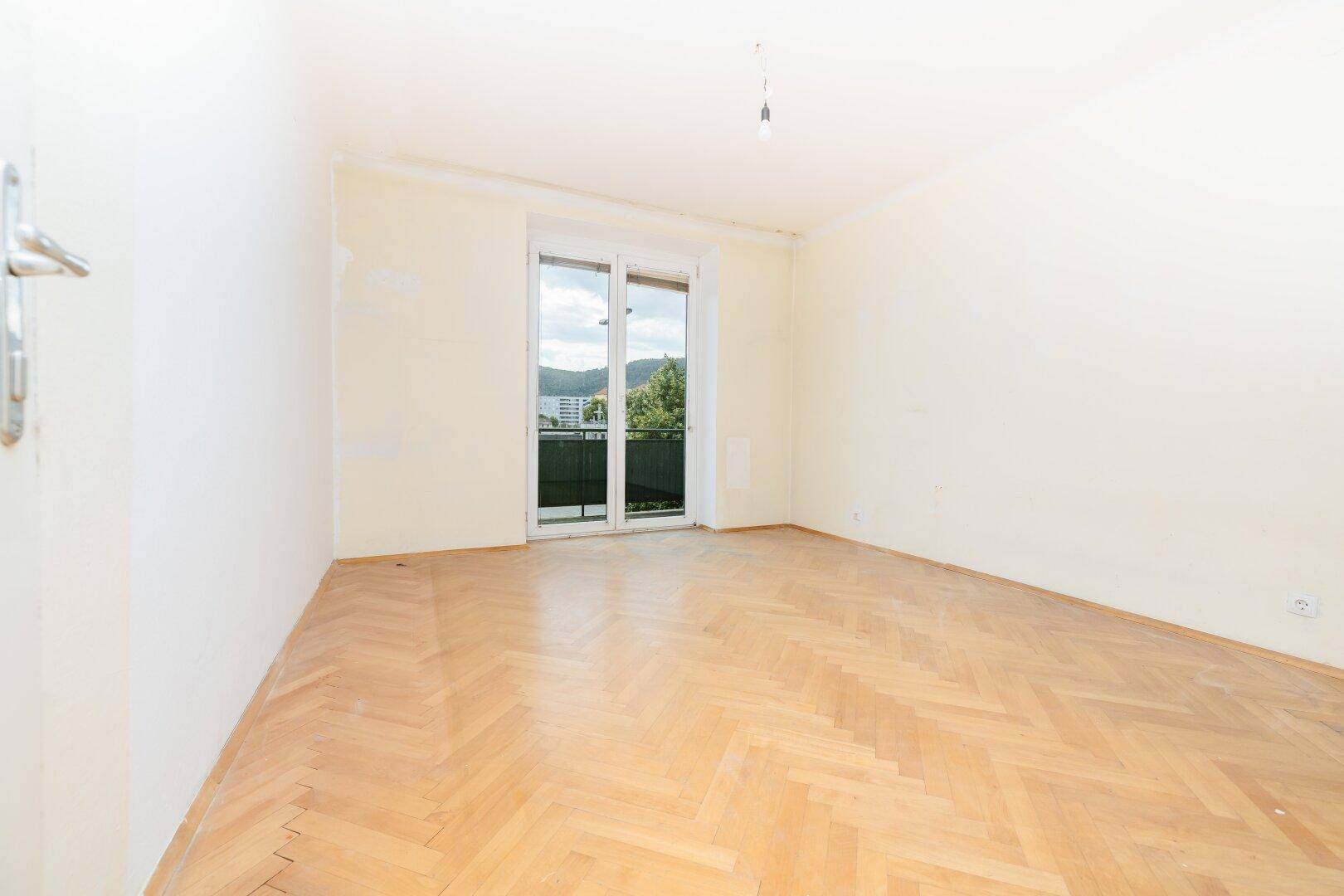 Helles Zimmer mit Fischgrätparkettboden und Zugang zu einem Balkon mit Aussicht.